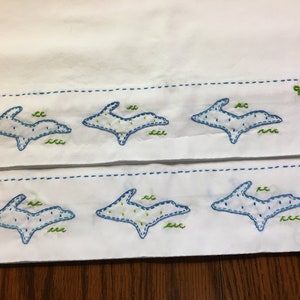 Op de afbeelding: Witte stof met twee rijen blauwe geborduurde contouren van de staat Michigan. De bovenste rij heeft het nummer 906 geborduurd in de rechterbovenhoek. De onderste rij heeft het woord "UP" geborduurd in de linkerbenedenhoek.