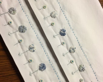 May include: White fabric with embroidered floral accents. The design features small, circular fabric flowers in shades of blue and gray, connected by gray thread to small buttons. Light blue stitching forms vertical lines.