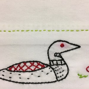 May include: White fabric with embroidered loon design. The loon is white with black outlines, a red eye, and a red and black patterned back. Green stitched line above the loon and green grass.