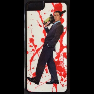 以下が含まれることがあります： 黒い縁取りのある白いiPhoneケース。ケースには、スーツを着た男性がチェーンソーを持っているグラフィックが描かれており、背景には赤い塗料の飛沫が描かれている。
