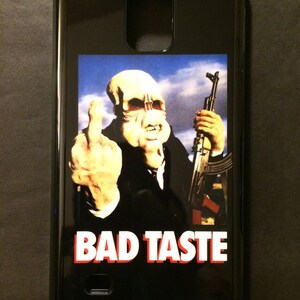 Könnte beinhalten: Schwarze Handyhülle mit einer Grafik einer Kreatur aus dem Film "Bad Taste", die den Mittelfinger zeigt und ein Gewehr hält. Der Text "Bad Taste" ist in roten und weißen Buchstaben gedruckt.