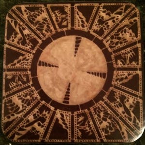 May include: A brown and black wooden puzzle box with a circular design in the center. The box has a series of interlocking pieces that must be manipulated in a specific order to open it.