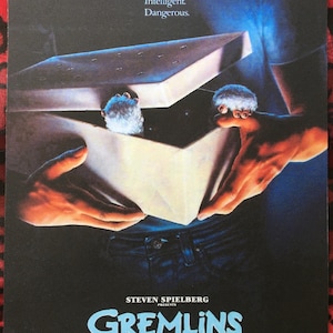 May include: A movie poster for the film Gremlins, featuring a person holding a box with two furry creatures inside. The text on the poster reads "Cute. Clever. Mischievous. Intelligent. Dangerous." and "Steven Spielberg Presents Gremlins".