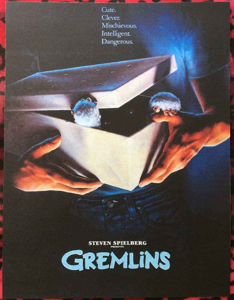 May include: A movie poster for the film Gremlins, featuring a person holding a box with two furry creatures inside. The text on the poster reads "Cute. Clever. Mischievous. Intelligent. Dangerous." and "Steven Spielberg Presents Gremlins".