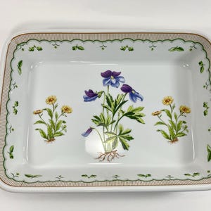 May include: A rectangular white ceramic baking dish featuring a central botanical illustration of purple pansies flanked by two smaller yellow flower clusters. The rim is decorated with a scalloped green border and a thin brown pinstripe pattern.