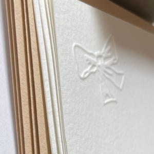 May include: A stack of white card stock with a subtle embossed bow design. The card stock is layered on top of brown card stock.