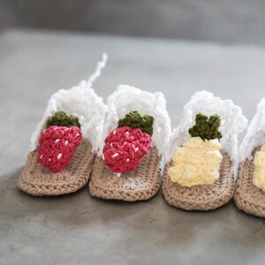 May include: Four pairs of crocheted baby sandals with fruit designs. Two pairs feature red strawberries and two pairs feature yellow pineapples.