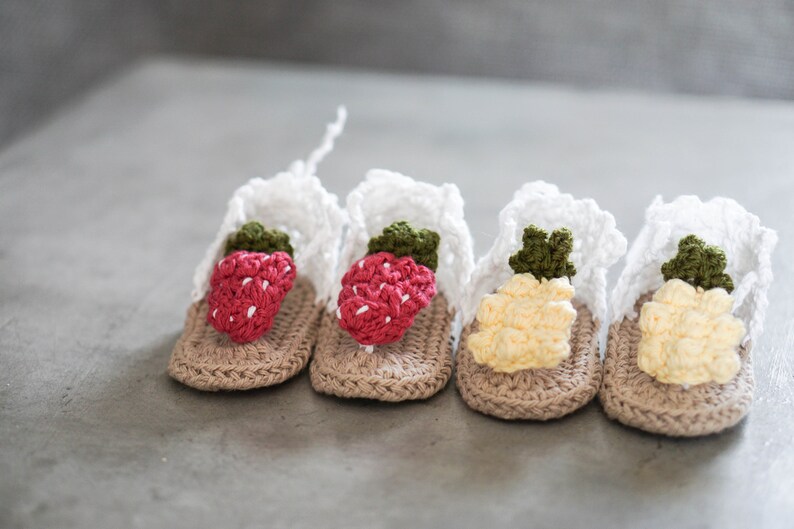 May include: Four pairs of crocheted baby sandals with fruit designs. Two pairs feature red strawberries and two pairs feature yellow pineapples.