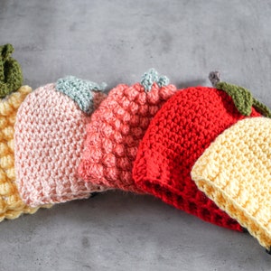 May include: Five crocheted hats in the shape of fruit. The hats are yellow, pink, orange, red, and yellow. The hats are arranged in a row on a gray surface.