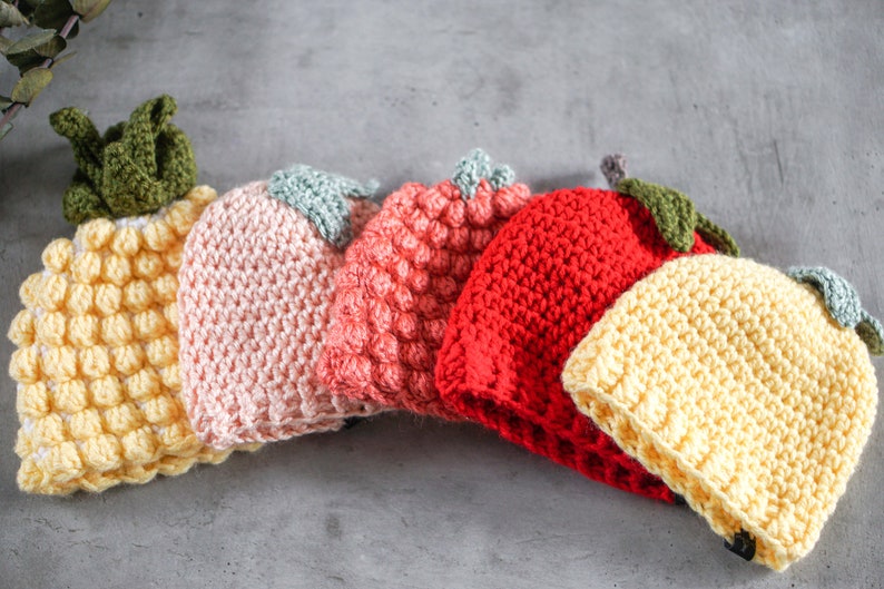 May include: Five crocheted hats in the shape of fruit. The hats are yellow, pink, orange, red, and yellow. The hats are arranged in a row on a gray surface.