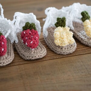 May include: Three pairs of crocheted baby sandals with different fruit designs. The sandals are beige with white straps and feature a strawberry, a pineapple, and a lemon.