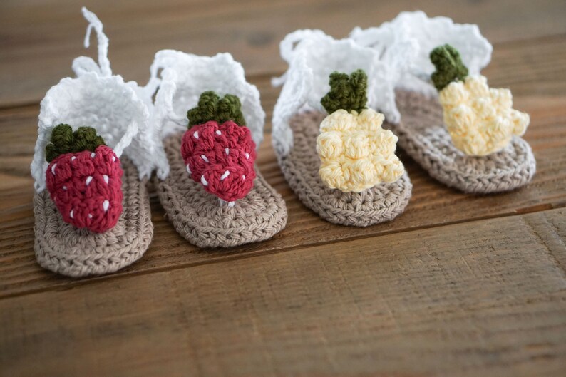 May include: Three pairs of crocheted baby sandals with different fruit designs. The sandals are beige with white straps and feature a strawberry, a pineapple, and a lemon.
