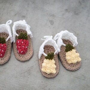 May include: Four crocheted baby sandals with white straps and fruit designs. Two sandals have red strawberry designs and two sandals have yellow pineapple designs.