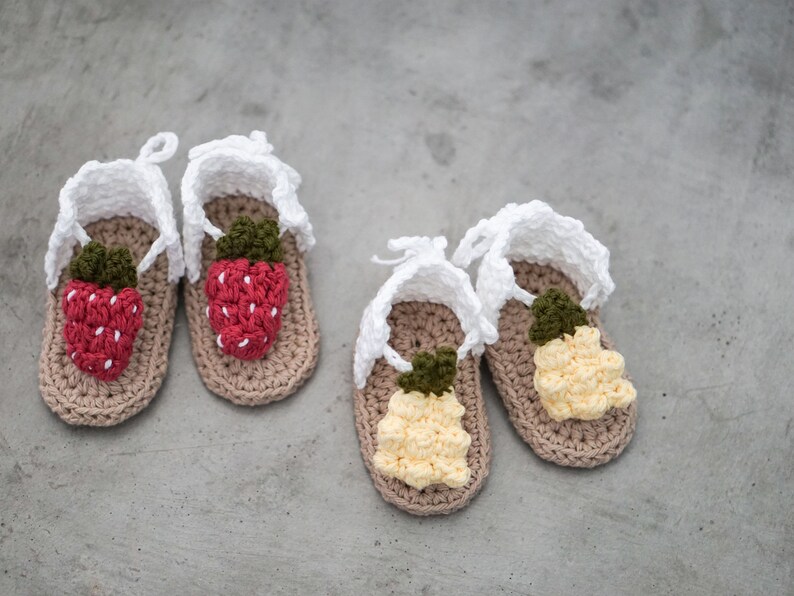 May include: Four crocheted baby sandals with white straps and fruit designs. Two sandals have red strawberry designs and two sandals have yellow pineapple designs.