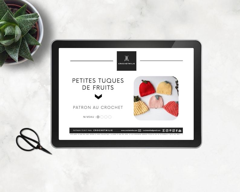 May include: A digital screen displaying a crochet pattern for making fruit-themed hats. The pattern is titled "Petites Tuques de Fruits" and includes a photo of five crocheted hats in the shape of strawberries, lemons, and oranges.