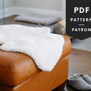 May include: A white faux fur rug is draped over a brown leather ottoman. The rug has a wavy edge and is soft and fluffy. The ottoman is in a living room setting with a wooden floor.