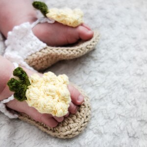 May include: A pair of crocheted baby sandals with yellow flower accents. The sandals are made with a beige and white yarn and have a delicate, handmade look.