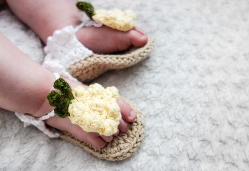May include: A pair of crocheted baby sandals with yellow flower accents. The sandals are made with a beige and white yarn and have a delicate, handmade look.