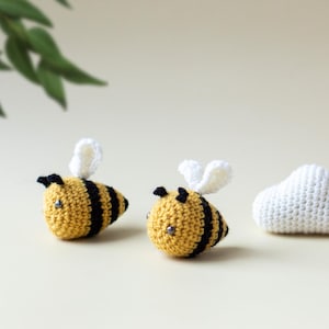May include: Two crocheted bumblebees with black and yellow stripes and white wings. The bees are sitting on a light beige surface. A white crocheted cloud is in the background.