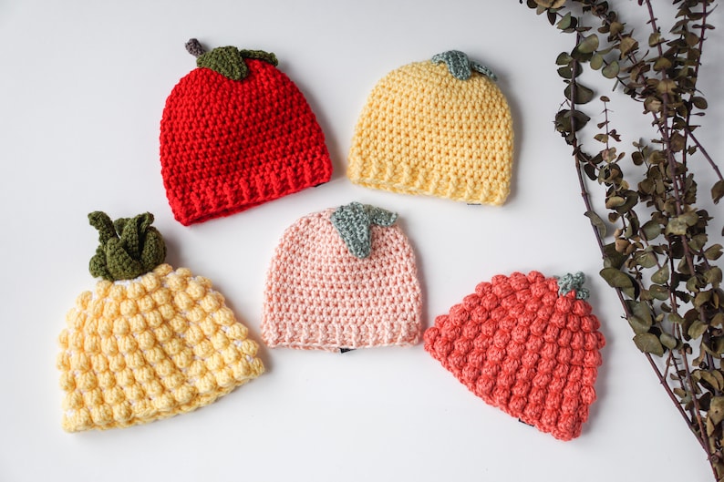 May include: Five crocheted hats in different fruit shapes and colors. The hats are yellow, red, pink, and orange. The hats have green crocheted leaves on top.