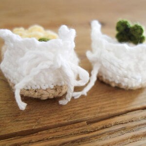 May include: A pair of white crocheted baby booties with a brown woven sole. One bootie has a yellow and white flower embellishment, while the other has a green flower embellishment.