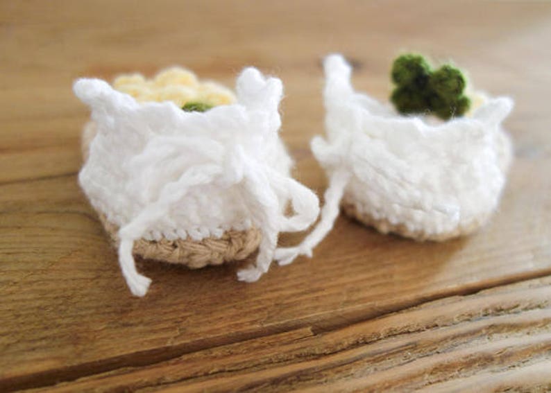 May include: A pair of white crocheted baby booties with a brown woven sole. One bootie has a yellow and white flower embellishment, while the other has a green flower embellishment.