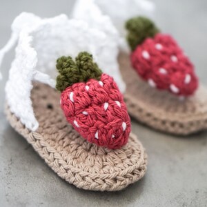 May include: A pair of crocheted baby sandals featuring a strawberry design. The sandals are beige with a white strap and a red strawberry with green leaves.