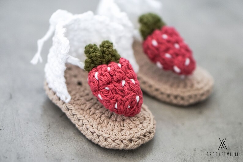 May include: A pair of crocheted baby sandals featuring a strawberry design. The sandals are beige with a white strap and a red strawberry with green leaves.