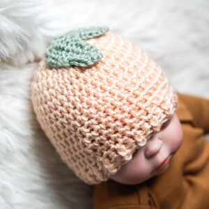 May include: A peach-colored knitted baby hat with a green crocheted leaf detail on top.