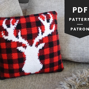 May include: A red and black plaid crochet pillow with a white deer silhouette. The pillow is on a gray couch with the text "PDF PATTERN PATRON" in black on a white background.