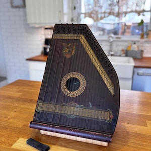 May include: A vintage, dark-stained wooden zither with a decorative eagle emblem and a circular sound hole. The instrument has strings, a tuning key, and a bridge. The zither rests on a wooden surface.