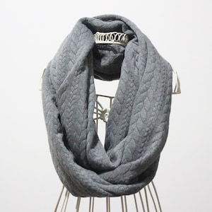 May include: A grey infinity scarf with a cable knit design. The scarf is displayed on a white wire mannequin. The fabric looks soft and warm, ideal for colder weather. The scarf's neutral colour makes it versatile.