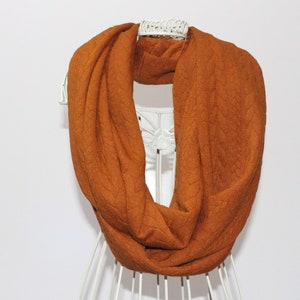 May include: An orange infinity scarf with a textured, quilted pattern. The scarf is displayed on a white mannequin, showcasing its circular design. The warm colour and soft texture suggest a cosy accessory.