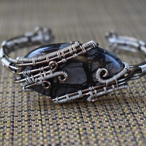 May include: A handcrafted silver wire cuff bracelet featuring a large, oval-shaped black stone with intricate patterns. The stone is securely wrapped with detailed wirework, showcasing a unique, artistic design. The bracelet is displayed on a textured, neutral-toned surface.