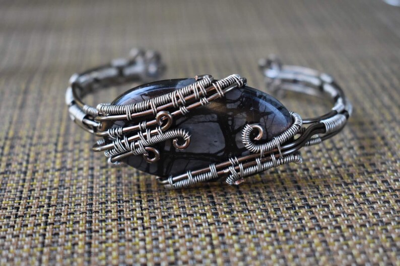 May include: A handcrafted silver wire cuff bracelet featuring a large, oval-shaped black stone with intricate patterns. The stone is securely wrapped with detailed wirework, showcasing a unique, artistic design. The bracelet is displayed on a textured, neutral-toned surface.