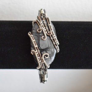 May include: A handcrafted pendant featuring a polished, oval-shaped stone with a gray and black pattern. The stone is intricately wrapped with silver-colored wire, creating a unique and artistic design. The pendant is suspended on a black background.
