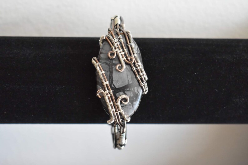 May include: A handcrafted pendant featuring a polished, oval-shaped stone with a gray and black pattern. The stone is intricately wrapped with silver-colored wire, creating a unique and artistic design. The pendant is suspended on a black background.