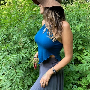 May include: A brown floppy hat with a black band is worn with a teal halter top and a gray skirt. The woman is standing in front of green foliage. A silver bracelet is on the wrist.