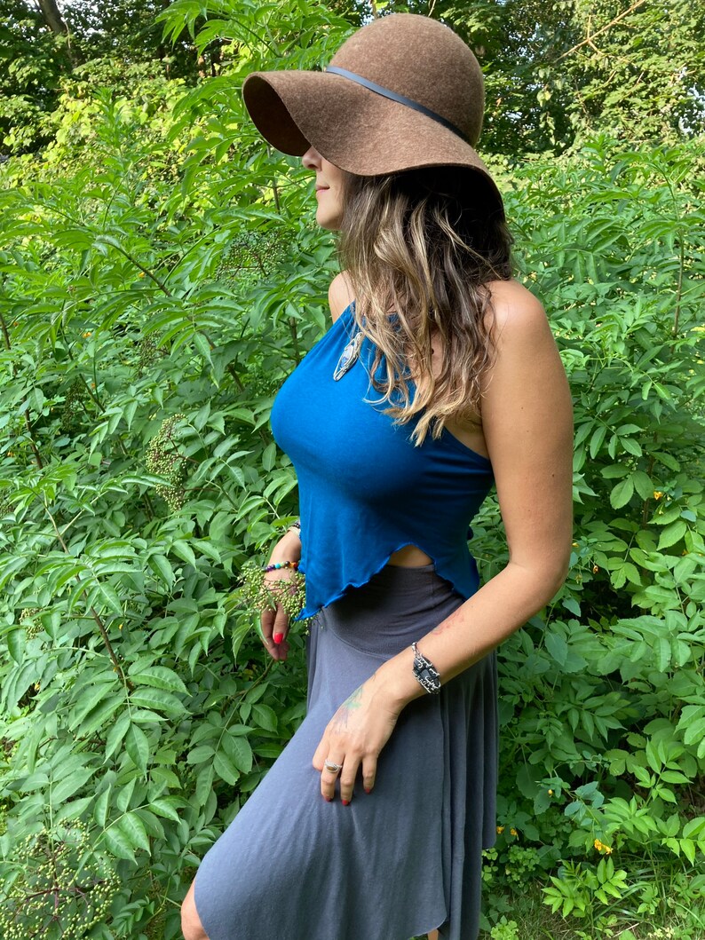 May include: A brown floppy hat with a black band is worn with a teal halter top and a gray skirt. The woman is standing in front of green foliage. A silver bracelet is on the wrist.