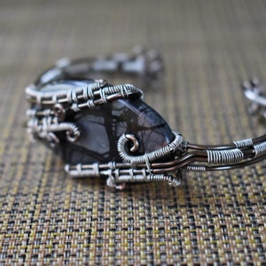 May include: A handcrafted cuff bracelet featuring a dark, patterned stone set within a silver-colored wire frame. The wire is intricately woven around the stone, creating a unique, artistic design. The bracelet is a piece of jewelry.