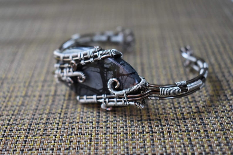 May include: A handcrafted cuff bracelet featuring a dark, patterned stone set within a silver-colored wire frame. The wire is intricately woven around the stone, creating a unique, artistic design. The bracelet is a piece of jewelry.
