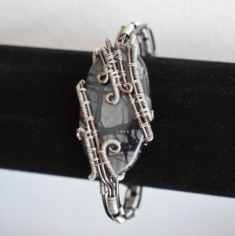 May include: A handcrafted bracelet featuring a dark gray, oval-shaped stone with a unique pattern. The stone is intricately wrapped with silver-colored wire, creating a decorative and artistic design. The bracelet is displayed against a black background.