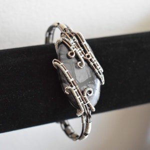 May include: A handcrafted bracelet featuring a dark gray stone with a unique pattern, encased in intricate silver-colored wire. The wirework forms a decorative cage around the stone, creating a stylish accessory. The bracelet is displayed on a black surface.