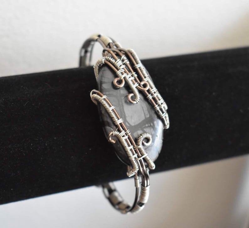 May include: A handcrafted bracelet featuring a dark gray stone with a unique pattern, encased in intricate silver-colored wire. The wirework forms a decorative cage around the stone, creating a stylish accessory. The bracelet is displayed on a black surface.