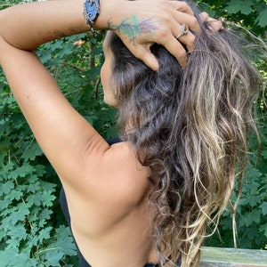 May include: A silver ring with a gold-colored center and a wire-wrapped bracelet with a dark blue stone are visible. The person has long, wavy hair with blonde highlights and a tattoo of a tree on their arm. The person is wearing a black top.