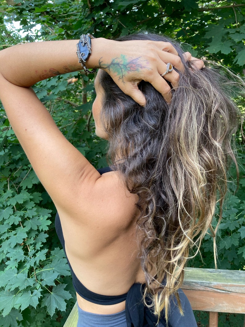 May include: A silver ring with a gold-colored center and a wire-wrapped bracelet with a dark blue stone are visible. The person has long, wavy hair with blonde highlights and a tattoo of a tree on their arm. The person is wearing a black top.