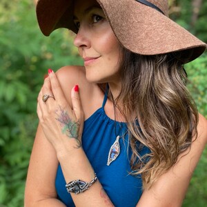 May include: A woman wearing a brown floppy hat and a blue halter top. She is wearing a silver bracelet with a black stone, a silver ring, and a pendant necklace with a wire-wrapped stone. Her nails are painted red.