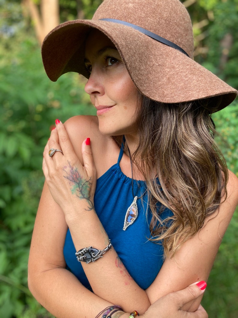 May include: A woman wearing a brown floppy hat and a blue halter top. She is wearing a silver bracelet with a black stone, a silver ring, and a pendant necklace with a wire-wrapped stone. Her nails are painted red.