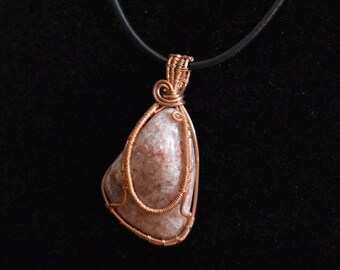 May include: A teardrop-shaped sunstone pendant featuring a polished, speckled peach and tan stone encased in intricate, hand-wrapped copper wire. The pendant hangs from a simple black cord, showcasing a detailed wire-coiled bail at the top.