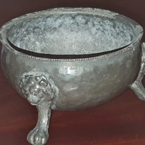 May include: A silver bowl with a lion head design on each of the three legs. The bowl has a slightly oval shape and a smooth, slightly textured surface.